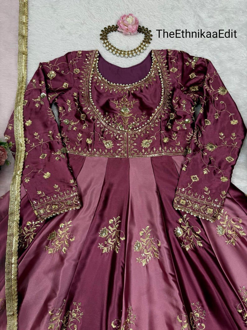 May include: A burgundy and mauve-colored formal dress with long sleeves. The dress features intricate gold embroidery on the bodice and sleeves, with a flowing skirt. A gold necklace with a pink flower and the text "TheEthnikaaEdit" are also visible.