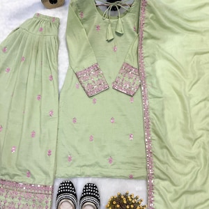 May include: A light green embroidered three-piece suit with matching pants and a scarf. The suit features pink floral embroidery and mirror work on the cuffs and pants. Black and white beaded flats are placed in front of the suit.