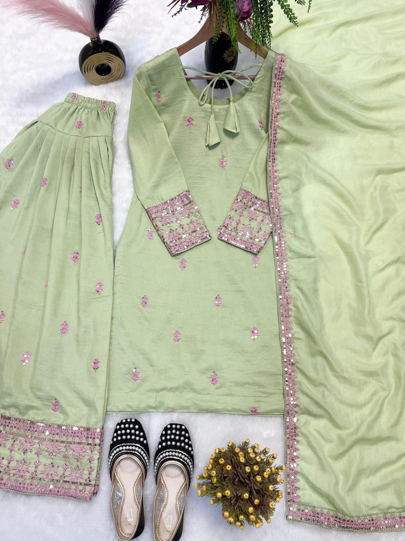 May include: A light green embroidered three-piece suit with matching pants and a scarf. The suit features pink floral embroidery and mirror work on the cuffs and pants. Black and white beaded flats are placed in front of the suit.