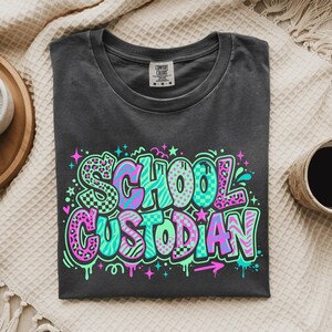 May include: A charcoal gray crew neck t-shirt featuring the text SCHOOL CUSTODIAN in a vibrant, multi-colored graffiti-style font. The letters incorporate neon pink, teal, and purple patterns including leopard print, checkers, and stripes.