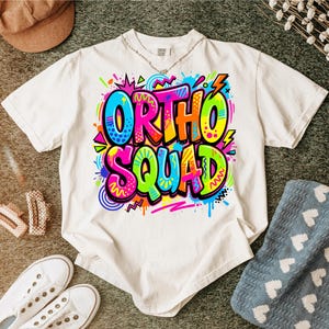 Może przedstawiać: Biały t-shirt z krótkim rękawem z żywym, wielokolorowym nadrukiem graficznym na klatce piersiowej. Wzór przedstawia napis "ORTHO SQUAD" pogrubioną czcionką inspirowaną graffiti z jasnymi neonowymi akcentami w kolorze różowym, niebieskim, żółtym i pomarańczowym.