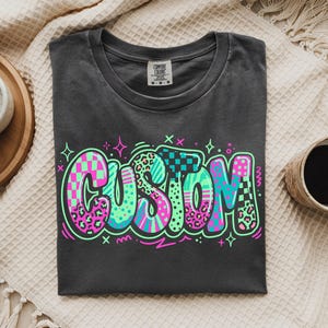 Op de afbeelding: Een antracietgrijs t-shirt met het woord CUSTOM in grote, levendige graffiti-stijl letters. De letters zijn gevuld met neonroze, turquoise en groene patronen, waaronder luipaardprint, ruitjes en strepen, omlijnd in fel neongroen.