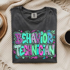 May include: A charcoal gray crew neck t-shirt featuring the text "BEHAVIOR TECHNICIAN" in a vibrant, multi-colored graphic design. The letters incorporate patterns including pink leopard print, teal checkerboard, and solid bright green.