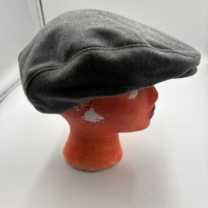 May include: A gray, flat cap made of a soft material. The hat is displayed on an orange mannequin head. The cap has a rounded crown and a small, stiff brim. The hat is a solid, neutral color.
