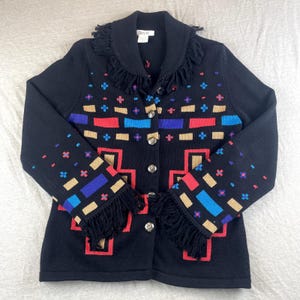 Könnte beinhalten: Schwarze Strickjacke mit farbenfrohem geometrischen Muster in Rot, Blau, Beige und Lila. Der Kragen und die Manschetten haben Fransen, und die Knöpfe sind silberfarben. Der Markenname "Bleyle" ist auf einem Etikett zu sehen.