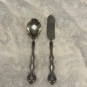 Puede incluir: Juego de cubiertos que incluye una cuchara de servir y un cuchillo de mantequilla. La cuchara tiene un cuenco festoneado y un mango ornamentado. El cuchillo de mantequilla tiene una hoja plana y un mango a juego. Ambas piezas son plateadas y tienen un diseño floral detallado.