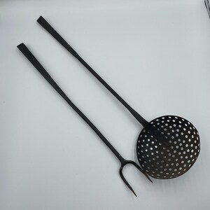 Op de afbeelding: Een paar keukengerei met lange stelen gemaakt van donker metaal. Het ene gereedschap is een vork met twee tanden en een slanke steel, het andere is een ronde schuimspaan met een geperforeerde kom en een bijpassende lange, slanke steel.