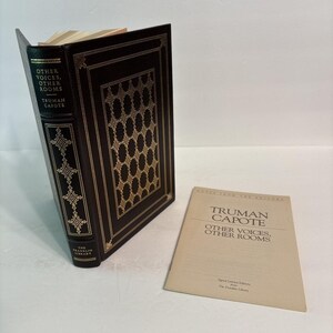 Könnte beinhalten: Ein in schwarzes Leder gebundenes Buch mit dem Titel Other Voices, Other Rooms von Truman Capote mit Goldfolienverzierungen auf dem Einband und Buchrücken, begleitet von einer beigen Papierbroschüre mit gleichem Titel und Autor.