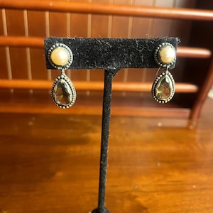 Puede incluir: Un par de pendientes colgantes exhibidos en un soporte de terciopelo negro. Cada pendiente presenta una perla redonda de color crema sobre una piedra preciosa en forma de lágrima, de color ahumado. Los pendientes tienen un borde con cuentas plateadas.