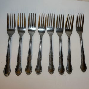 Peut inclure: Un ensemble de huit fourchettes en métal argenté disposées en rangée. Chaque fourchette possède quatre dents et un manche orné d'un motif de volutes en relief sur les bords, se terminant par une base arrondie avec un détail floral.