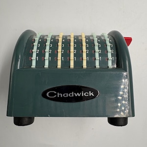 Pode incluir: Uma máquina de somar manual Chadwick verde-escura com uma série de colunas numéricas deslizantes verticais com abas de plástico verde-claras e creme. O dispositivo tem uma placa preta com o texto Chadwick e pés de borracha pretos.