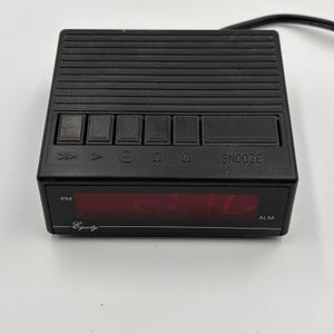 May include: A black rectangular digital alarm clock with a red LED display. The top features horizontal ridges and a row of control buttons labeled with icons and the word SNOOZE. The front display shows PM and ALM indicators, with the brand name Equity printed in white script on the lower left corner.