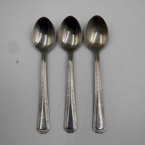 May include: Three silver-toned metal spoons arranged side by side. Each spoon features a smooth, oval-shaped bowl and a long, slender handle decorated with a series of vertical, parallel etched lines that run down the center of the stem.