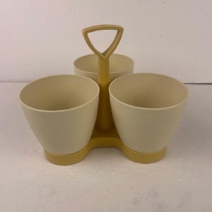 Puede incluir: Un especiero vintage de color crema con tres cuencos redondeados y un asa amarilla. El especiero es de plástico y tiene una base amarilla. Los cuencos están dispuestos en forma triangular, para sal, pimienta y otras especias.