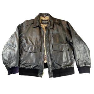 May include: A black leather bomber jacket with two front pockets, a collared neckline, and a ribbed hem and cuffs. The jacket has a map-like patterned lining and a button closure. The sleeves have a slight puff at the shoulders.