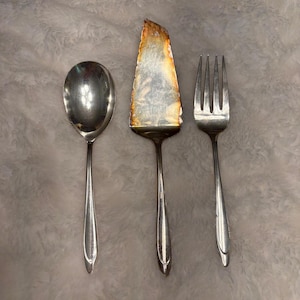 May include: Silverware set including a serving spoon, cake server, and fork. The cake server has a tarnished blade. The spoon and fork have long, slender handles. The set is likely vintage and suitable for serving at a formal dinner.