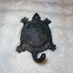 May include: A dark bronze-colored turtle figurine with a textured shell and detailed legs and head. The turtle has a curved tail and is displayed on a white, fluffy surface. The figurine is a decorative item.