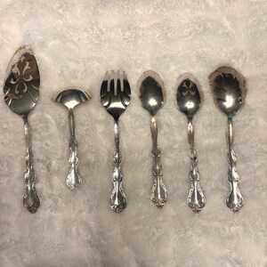 May include: A set of six antique silver serving utensils. The set includes a pie server, a small ladle, a serving fork, and three different serving spoons. Each piece has a decorative handle and ornate detailing.