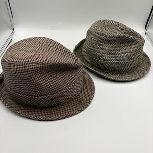 May include: Two tweed hats in shades of brown and green. One hat has a flat cap style with a houndstooth pattern, while the other is a fedora with a plaid design. Both hats have a brim and are made of a textured fabric.