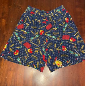May include: Navy blue shorts with a white polka dot pattern and a repeating print of beauty supplies. The print includes combs, lipsticks, keys, and other items in red, yellow, and green.