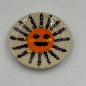 Könnte beinhalten: Eine runde, cremefarbene Keramikschale mit einem zentralen orangefarbenen Sonnengesicht mit schwarzen Augen und einem lächelnden Mund. Die Sonne ist von strahlenförmigen, violetten und gelben Bemalungen umgeben, die zum Rand hin verlaufen.