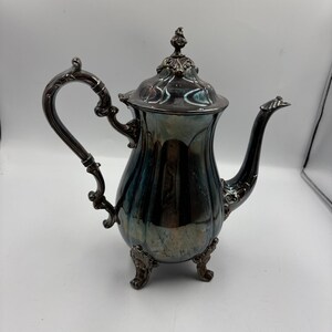 May include: A silver-toned metal teapot featuring a curved handle, a long spout, and a removable lid with a decorative finial. The body of the teapot has a fluted design and rests on three ornate, curved feet, showing a polished, reflective surface.