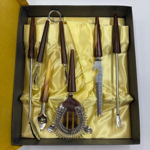 Può includere: Un set da bar per cocktail vintage in una scatola. Il set include uno strainer, un apribottiglie e vari strumenti da bar con manici marroni e beige. Gli strumenti sono disposti su un tessuto giallo satinato all'interno di una scatola scura.
