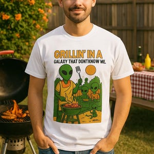 May include: White t-shirt with the text "Grillin' in a Galaxy That Don't Know Me." The shirt features a cartoon alien grilling food on a barbecue. The person is standing in a backyard with a grill and a picnic table.