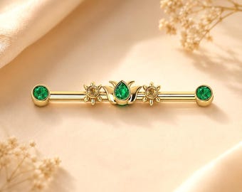 May include: A gold industrial barbell piercing with a central lotus flower design featuring a pear-cut green gemstone. The bar is accented by two smaller round yellow gemstones and two round green gemstones at the ends of the straight metal bar.