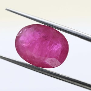 May include: An oval-shaped ruby gemstone, held by silver tweezers. The ruby has a deep pink-red colour and internal light reflections, highlighting its facets. The tweezers are against a white background. A gemstone for jewellery.