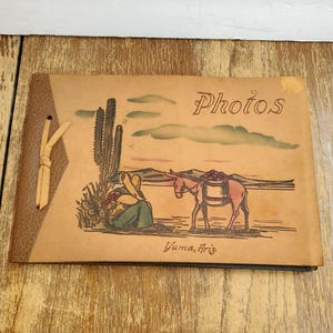 May include: A tan leather photo album with a tied spine and a desert scene illustration featuring a saguaro cactus, a person sitting, and a donkey carrying a basket. The cover includes the text Photos and Yuma, Ariz. in brown ink.
