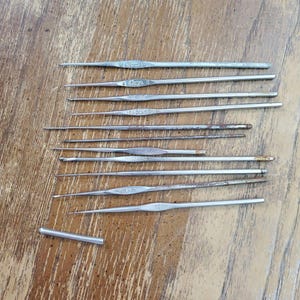May include: A collection of eleven silver-toned metal crochet hooks of varying lengths and thicknesses, some featuring embossed size markings on the flat center section, alongside one small detached metal cylinder piece.