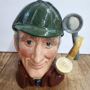 May include: A ceramic Sherlock Holmes head-shaped mug. The mug features a green cap, a brown coat, and a pipe. A magnifying glass is attached to the side. The face has a smiling expression with rosy cheeks.