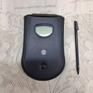 May include: A black Palm handheld device with a flip cover, a small screen, and a stylus. The device has a rounded shape with a circular button at the bottom and the Palm logo. The stylus is long and thin, with a pointed tip.