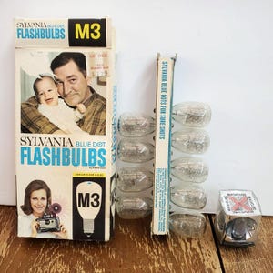 May include: Vintage Sylvania Blue Dot Flashbulbs boxes and flashbulbs. The boxes feature images of a man holding a baby and a woman holding a camera. The text on the boxes reads "SYLVANIA BLUE DOT FLASHBULBS" and "M3".