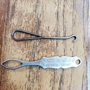 May include: A vintage metal button hook with a simple wire loop and a hook end, paired with a flat silver-toned metal tool featuring a diamond-shaped eye and the engraved text Sullivan-Cook Co. Men's Wear, both used for fastening clothing buttons.