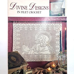 May include: A book titled "Divine Designs in Filet Crochet" by Vivian Floy Smith. The cover features a cream-colored filet crochet design of a religious figure. The image includes candles, a decorative book, and a small crochet sample.