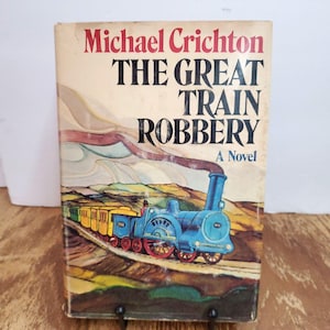 May include: Vintage paperback book titled "The Great Train Robbery" by Michael Crichton. The cover features a blue steam train with red wheels and yellow train cars. The title is in large black letters with the author's name in red.