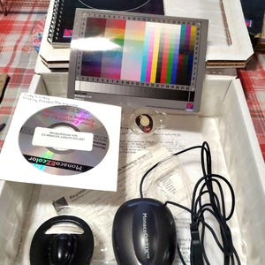 May include: A MonacoEZcolor kit featuring a spiral-bound user guide, a color calibration chart with a grid of colored squares, a software disc, a black MonacoOPTIX XR colorimeter device with a USB cable, and a circular black counterweight.