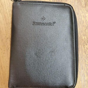May include: A rectangular dark brown zippered travel case with a textured surface. The brand name Samsonite is embossed in the center of the front panel, accompanied by the brand logo above the text. The case features a black zipper closure.