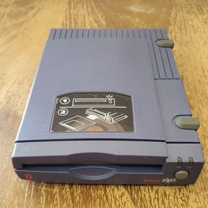 May include: A gray Iomega Zip drive, a vintage external storage device. The device has a rectangular shape with a sliding door and a power button. The front of the drive has the Iomega logo and the words "Iomega zip".