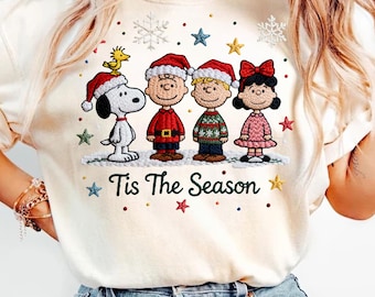 Könnte beinhalten: Cremefarbenes Sweatshirt mit einem gestickten Design von Snoopy, Woodstock, Charlie Brown, Linus und Lucy in Weihnachtskleidung. Der Schriftzug "Tis The Season" ist unter den Figuren mit Sternen und Schneeflocken gestickt.