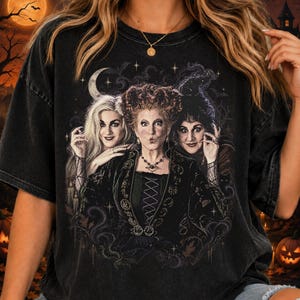 May include: A black acid-wash t-shirt featuring a graphic print of three witches with distinct hairstyles and dark clothing. The design includes a crescent moon, stars, and swirling patterns, set against a dark, atmospheric background with trees.