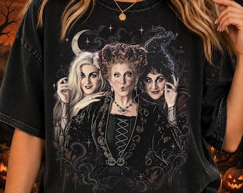 May include: A black acid-wash t-shirt featuring a graphic print of three witches with distinct hairstyles and dark clothing. The design includes a crescent moon, stars, and swirling patterns, set against a dark, atmospheric background with trees.