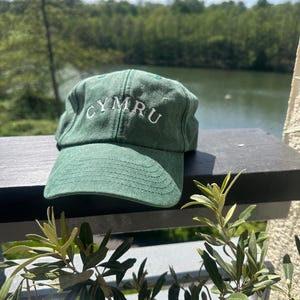 以下が含まれることがあります： 白い文字で「CYMRU」と刺繍された緑色の野球帽。帽子は、湖と木々の美しい背景を持つ暗い表面に置かれています。帽子には湾曲したつばがあります。