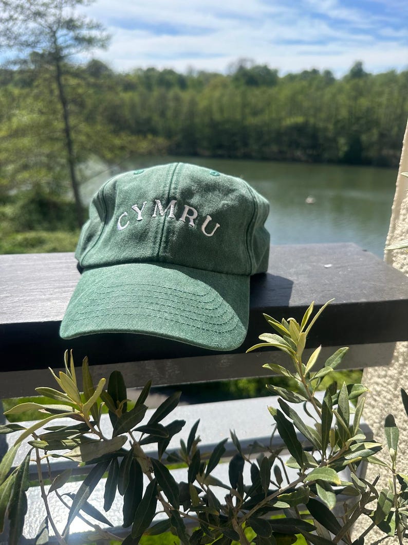 以下が含まれることがあります： 白い文字で「CYMRU」と刺繍された緑色の野球帽。帽子は、湖と木々の美しい背景を持つ暗い表面に置かれています。帽子には湾曲したつばがあります。