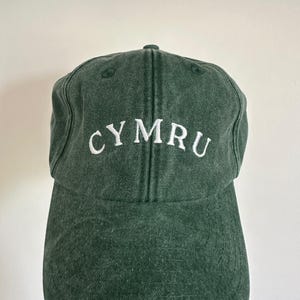 以下が含まれることがあります： 前面に白で「CYMRU」の文字が刺繍された濃い緑色の野球帽。 つばがカーブしており、上部が丸くなっています。 帽子は柔らかく、質感のある生地で作られています。