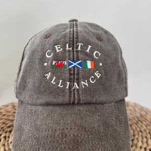 May include: A charcoal grey baseball cap featuring the embroidered text "CELTIC ALLIANCE" in white. Between the words are three small embroidered flags representing Wales, Scotland, and Ireland, arranged horizontally in a row.