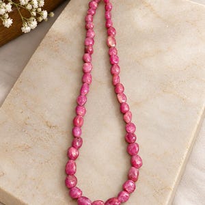 May include: A single strand necklace composed of numerous small, faceted, irregular pink gemstone beads. The beads are strung on a light-coloured cord, creating a continuous loop of vibrant, polished stones with varying shapes and sizes.