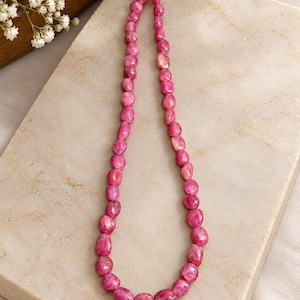 May include: A single strand necklace composed of numerous small, faceted, irregular pink gemstone beads. The beads are strung on a light-colored cord, creating a continuous loop of vibrant, polished stones with varying shapes and sizes.